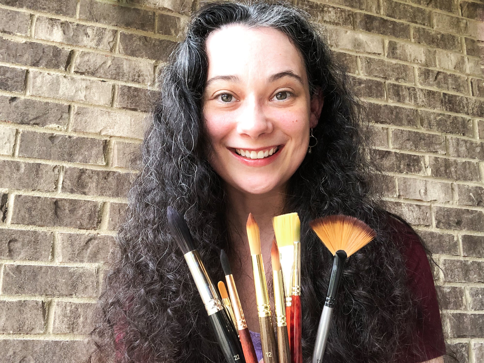 Artist Jennifer Diann of Lovely Joy Design in front of a brick wall holding various paintbrushes.